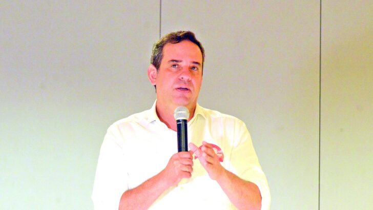 Homem de camisa branca fala ao microfone diante de uma parede clara
