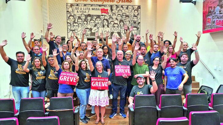 Grupo de bancários com camisetas pretas levanta as mãos em um auditório; alguns seguram cartazes com a frase Bancários em greve