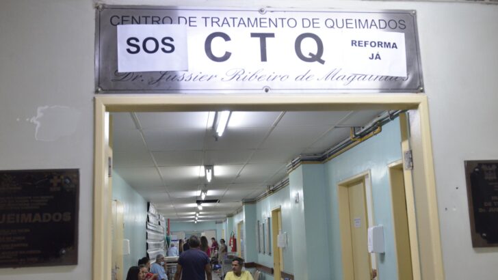 Entrada do Centro de Tratamento de Queimados com cartazes escritos SOS e Reforma já sobre a placa, e pacientes no corredor ao fundo