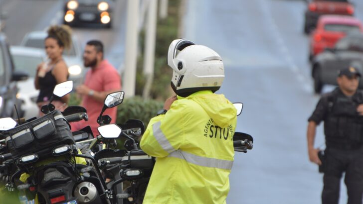 Agente de trânsito de capacete branco e jaqueta amarela com a inscrição STTU, ao lado de motocicletas, numa avenida movimentada