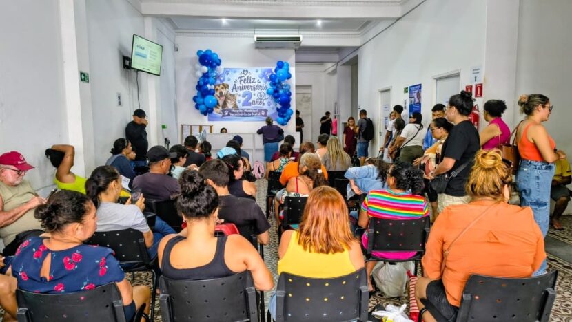 Sala de espera cheia de pessoas sentadas em cadeiras pretas, com painel de Feliz Aniversário 2 anos e balões azuis ao fundo