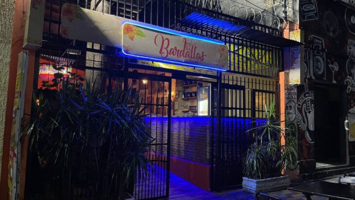 Fachada de bar à noite com letreiro iluminado escrito Bardallos, grades pretas e luz azul na entrada