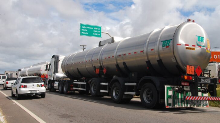Caminhão-tanque prateado com logotipo BR na beira de uma rodovia, com um carro branco e outros caminhões ao lado