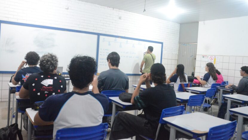 Estudantes sentados em carteiras azuis, de costas para a câmera, com um professor diante do quadro branco ao fundo