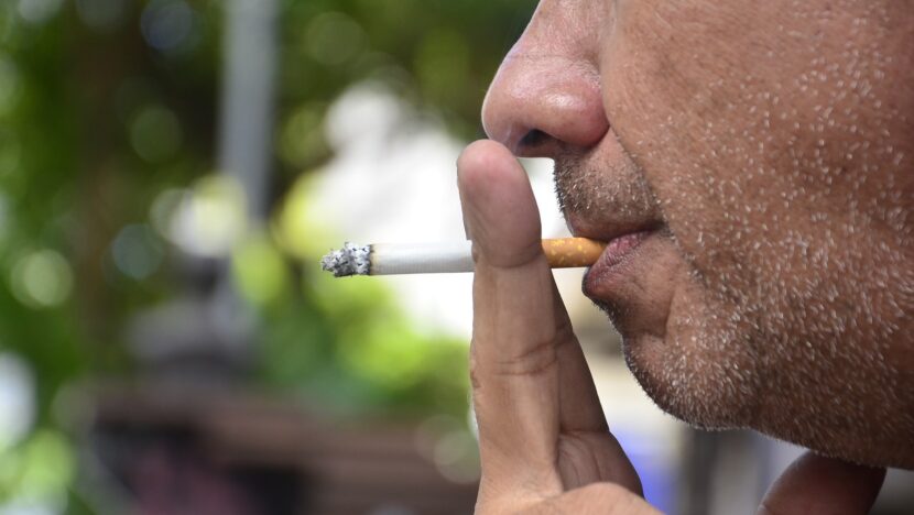 Close do rosto de um homem de barba grisalha por fazer, segurando um cigarro aceso junto à boca
