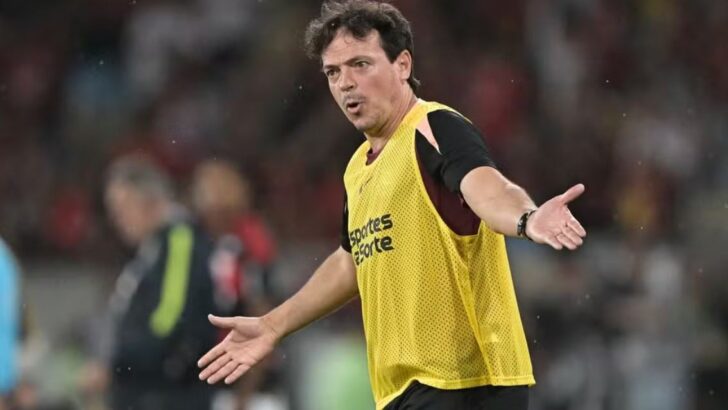Homem de cabelo castanho encaracolado, com colete amarelo sobre camisa preta, abre os braços à beira do campo