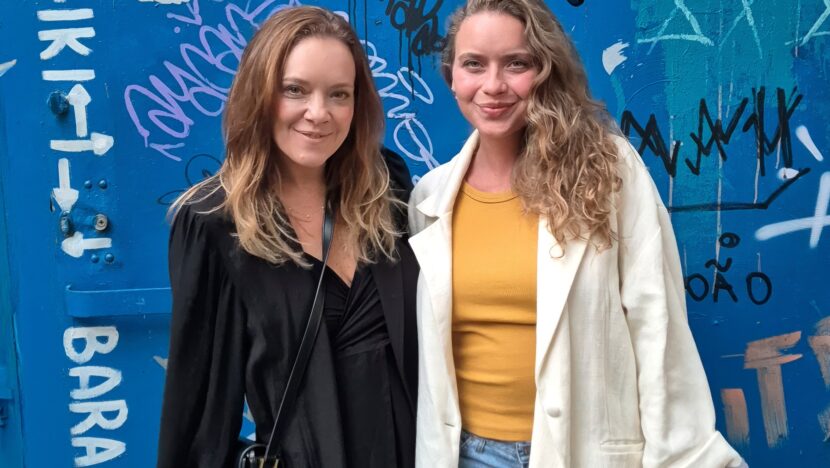 Duas mulheres lado a lado diante de uma parede azul grafitada; a da esquerda veste preto e a da direita, casaco claro sobre blusa amarela