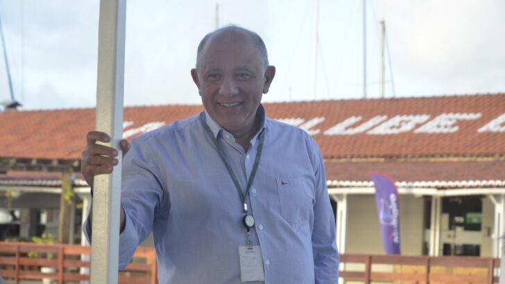 Homem de camisa social azul-clara e crachá sorri apoiado em um poste branco, com construção de telhado vermelho ao fundo