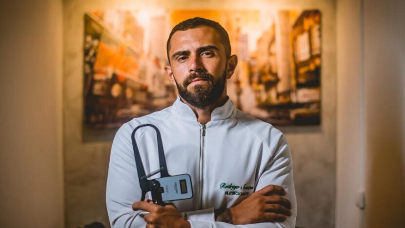 Homem de barba, com jaleco branco bordado com o nome Rodrigo Santos, de braços cruzados e segurando um adipômetro