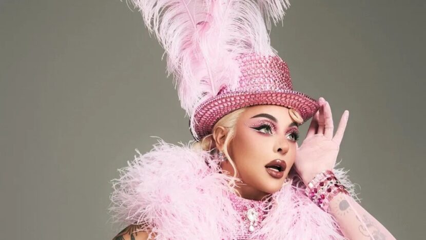 Cantora loira com cartola rosa de plumas, maquiagem marcante e boá de penas cor-de-rosa, com a mão junto ao rosto