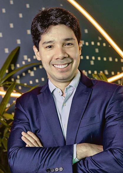 Homem de cabelo escuro, terno azul e camisa clara, sorrindo de braços cruzados diante de parede com iluminação decorativa