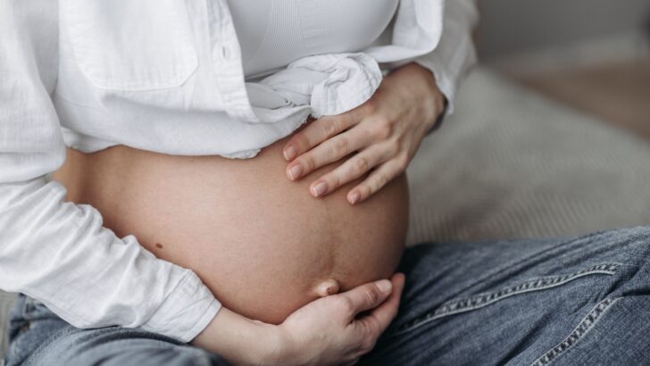 Gestante de camisa branca e calça jeans com as mãos sobre a barriga