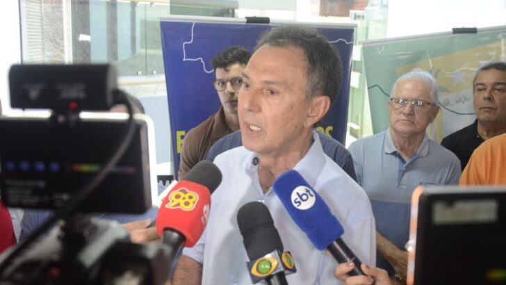 Homem de camisa social azul-clara fala a vários microfones de emissoras de rádio e TV, com outras pessoas ao fundo