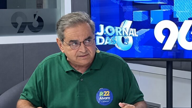 Homem de óculos e camisa polo verde com adesivo de campanha, sentado em estúdio de rádio com copos de água à frente