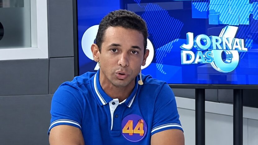 Homem de camisa polo azul com adesivo de campanha número 44, sentado em estúdio de rádio