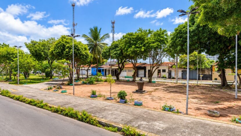 Praça com árvores, postes de iluminação, canteiros e busto ao centro, cercada por casas, sob céu azul
