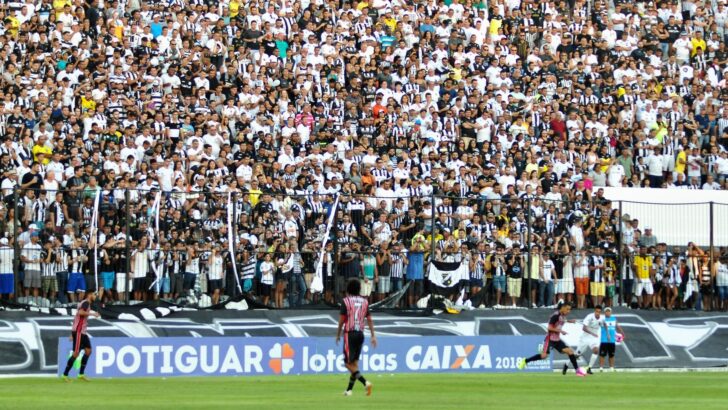 Arquibancada lotada de torcedores do ABC com camisas alvinegras no Frasqueirão, com jogadores no gramado à frente