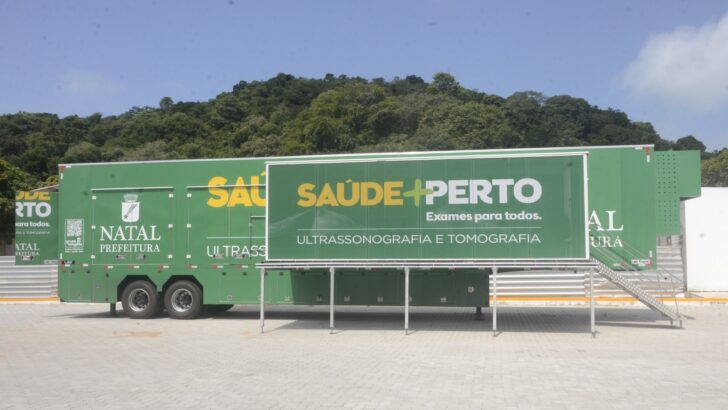 Carreta verde com a inscrição Saúde + Perto, ultrassonografia e tomografia, estacionada num pátio com morro ao fundo