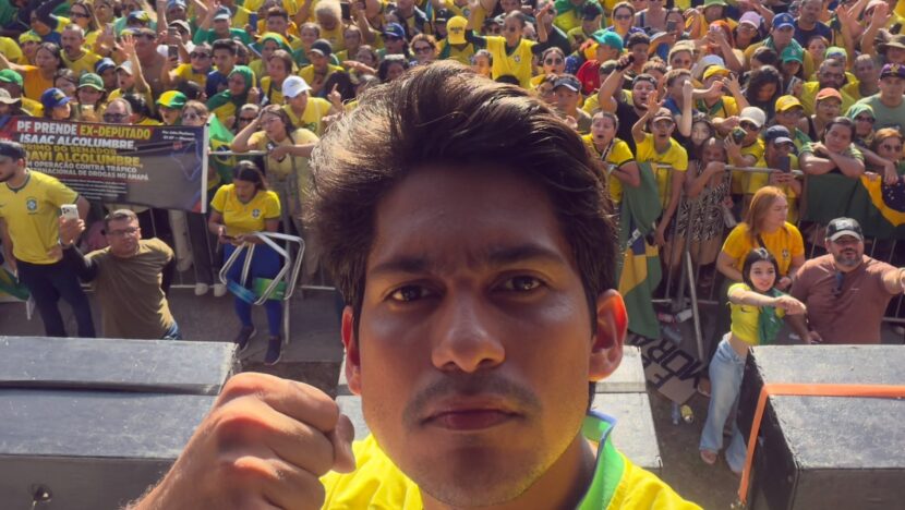 Homem jovem com camisa amarela da seleção brasileira ergue o punho em selfie diante de uma multidão vestida de verde e amarelo