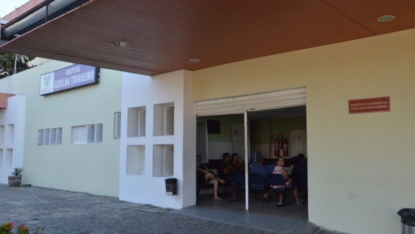 Fachada de hospital com cobertura de madeira e pacientes sentados na recepção, vistos pela porta de vidro aberta