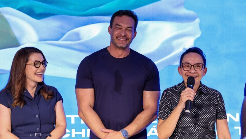 Homem de camiseta azul-escura em pé entre duas mulheres, uma delas falando ao microfone, diante de painel azul