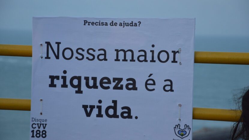 Cartaz com a frase Nossa maior riqueza é a vida e o número 188 do CVV, preso a uma grade amarela diante do mar