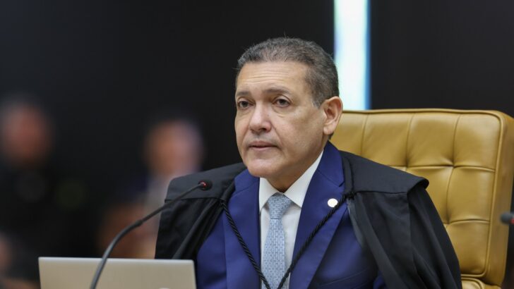 Homem de toga preta sobre terno azul, sentado em poltrona dourada diante de um notebook