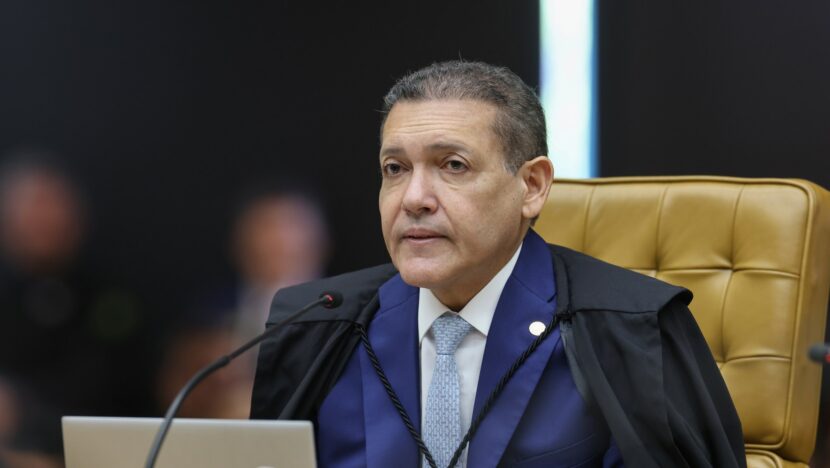 Homem de toga preta sobre terno azul, sentado em poltrona dourada diante de um notebook