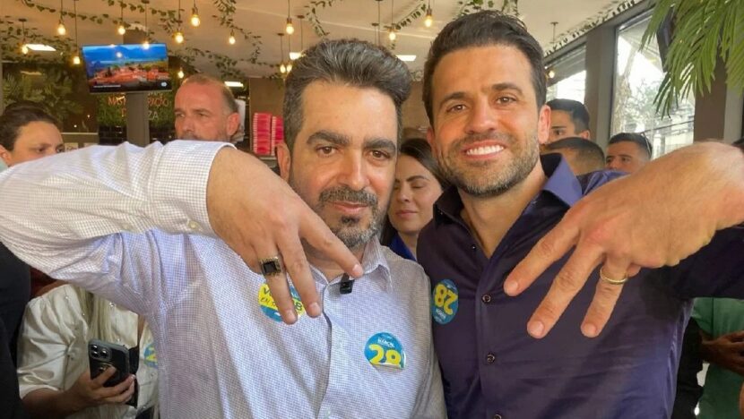 Dois homens sorridentes posam abraçados fazendo gestos com as mãos, em um ambiente cheio de pessoas