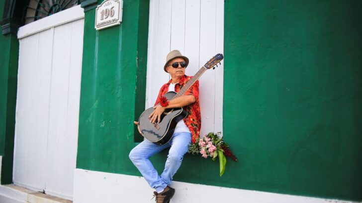 Homem de chapéu e camisa colorida toca violão sentado na soleira de uma casa com parede e portas verdes