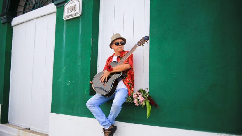 Homem de chapéu e camisa colorida toca violão sentado na soleira de uma casa com parede e portas verdes