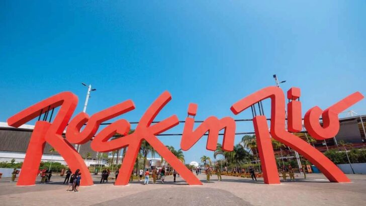 Letreiro vermelho gigante com as palavras Rock in Rio sob céu azul, com estruturas do festival ao fundo