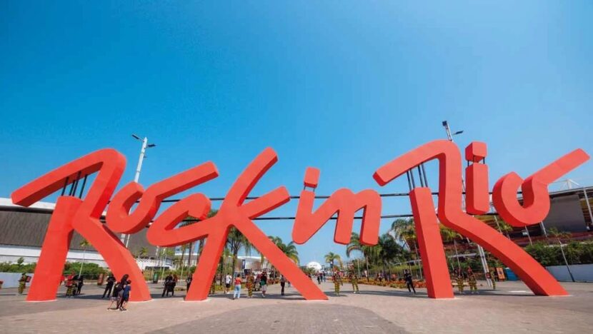 Letreiro vermelho gigante com as palavras Rock in Rio sob céu azul, com estruturas do festival ao fundo