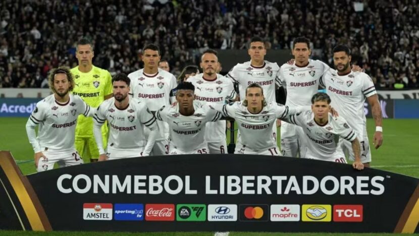 Jogadores do Fluminense de uniforme branco posam para foto oficial em campo, atrás de faixa da Conmebol Libertadores