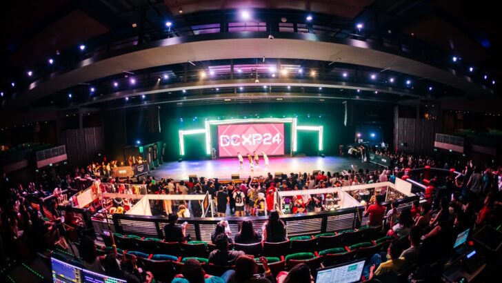 Arena com palco iluminado, telões e plateia acompanhando uma competição de jogos eletrônicos