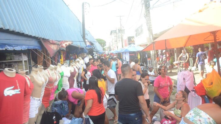 Rua de comércio popular cheia de pessoas, com barracas cobertas por lonas azuis e roupas expostas