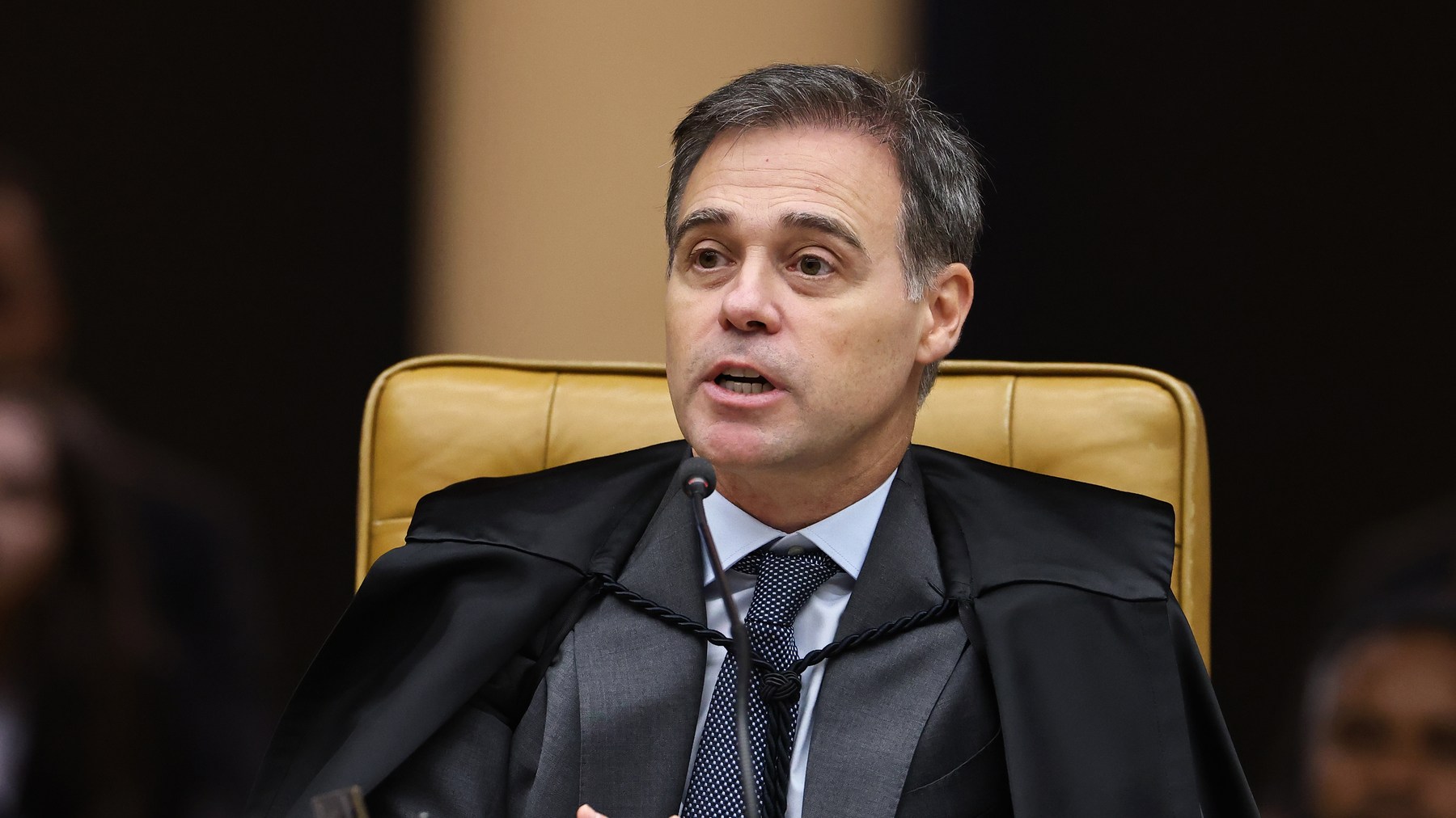 Ministro de terno escuro e gravata fala em sessão, sentado em poltrona alta de couro