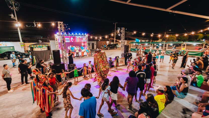 Público forma roda ao ar livre à noite diante de palco iluminado, ao lado de brincantes com trajes coloridos
