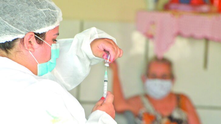Profissional de saúde com touca, máscara e avental prepara seringa diante de idosa sentada