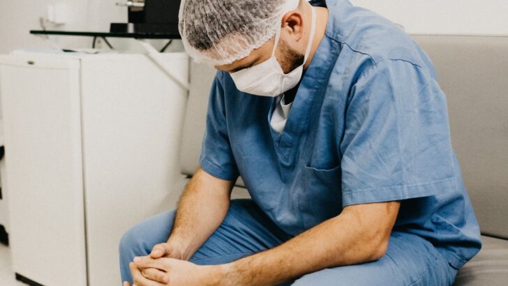 Profissional de saúde de uniforme, touca e máscara sentado com a cabeça baixa