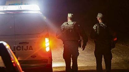 Dois homens de uniforme caminham à noite ao lado de viatura policial