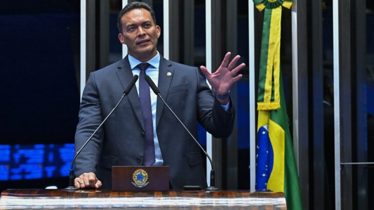 Senador de terno cinza discursa na tribuna do Senado, ao lado da bandeira do Brasil