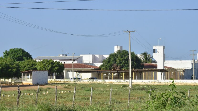 Muro e prédios de unidade prisional vistos de terreno com vegetação
