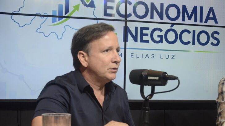 Homem de camisa escura fala ao microfone em estúdio com painel de economia e negócios