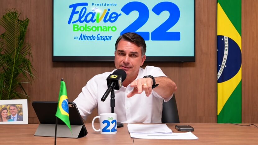 Homem de camisa branca fala ao microfone em mesa, diante de painel de campanha e bandeira do Brasil