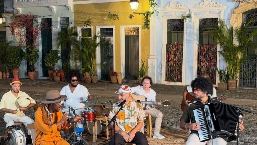 Grupo de músicos com sanfona, percussão e violão toca sentado em rua de casario colorido