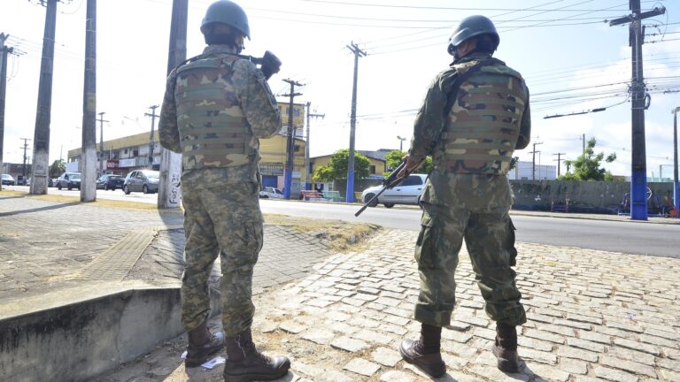 Dois agentes fardados, de capacete e colete, em pé numa rua, de costas para a câmera