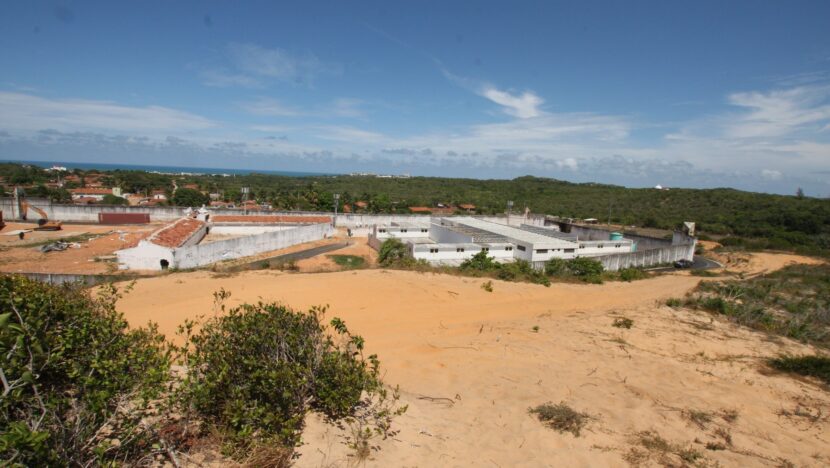 Vista de longe de um complexo prisional cercado por dunas e vegetação baixa, sob céu azul