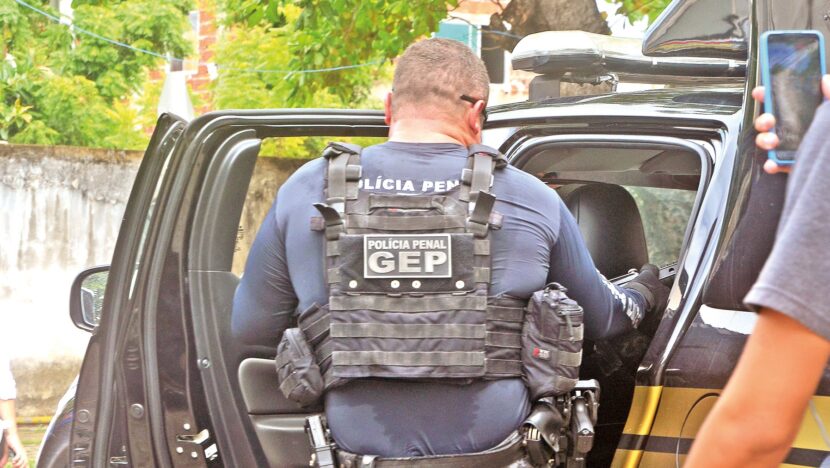 Policial penal de colete tático entra em viatura, de costas, com a palavra GEP escrita nas costas do colete