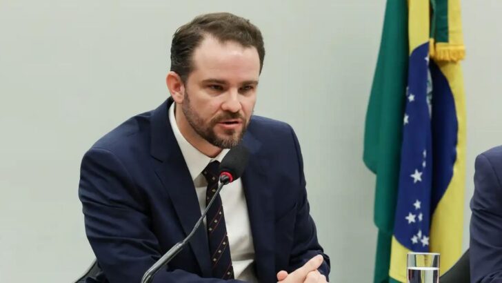 Homem de terno e gravata fala ao microfone durante evento, com expressão séria